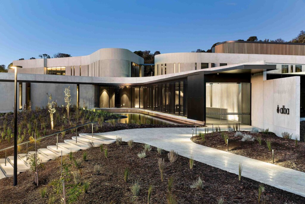 The stunning, curved architecture of the Alba retreat at dusk, with an illuminated stone pathway from Eigen Stones leading over a tranquil water feature.