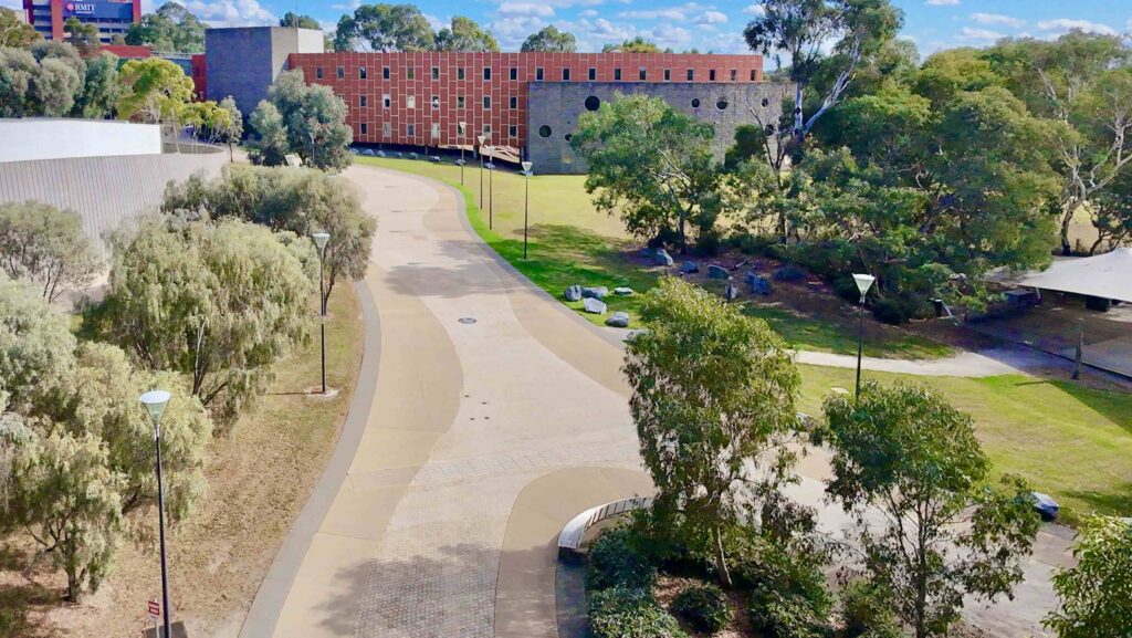  A wide, stone-paved plaza at Monash University, featuring a dark stone planter wall that creates a clean edge against a green lawn.