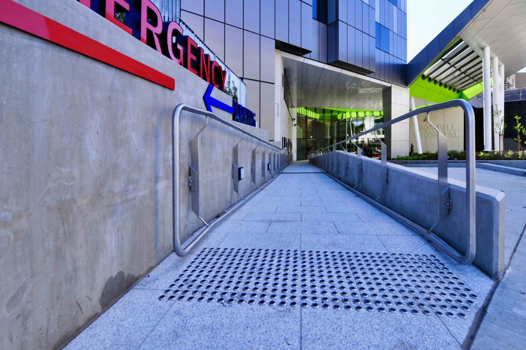 A sleek, stone-paved accessibility ramp with integrated tactile indicators leading to the Emergency entrance at Box Hill Hospital, with all stone supplied by Eigen Stones.